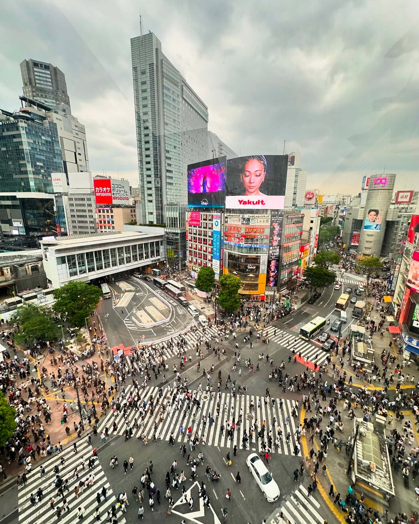 An aerial view of the Shibuya Crossing in Tokyo, showing a massive crowd of pedestrians crossing from multiple directions, surrounded by tall buildings adorned with vibrant neon signs and digital billboards at dusk.