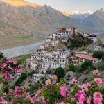 A panoramic view of a Buddhist monastery perched on a barren, rocky cliff in Spiti Valley, surrounded by towering, snow-dusted mountains under a dramatic sky.