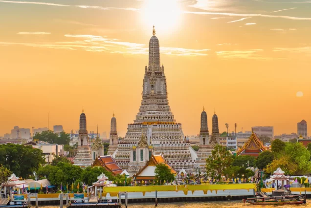 Wat Pho, a grand Buddhist temple in Bangkok, Thailand, seen from the Chao Phraya River, with its golden spires and ornate roofs reflecting on the water, and a cruise boat in the foreground.