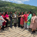A group of tourists, one wearing a traditional Bhutanese gho, stand on a hillside overlooking a lush valley with mountains and a dzong in the distance.