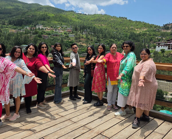 A group of tourists, one wearing a traditional Bhutanese gho, stand on a hillside overlooking a lush valley with mountains and a dzong in the distance.