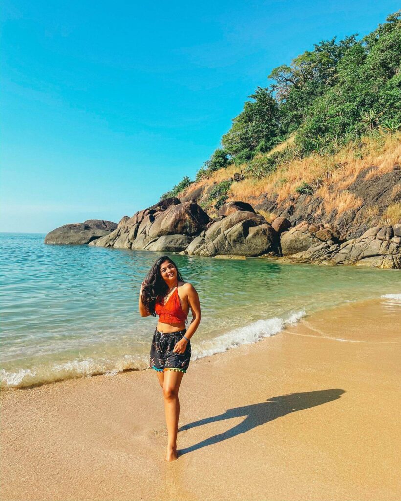 A scenic photograph of a quiet, sandy beach in Goa, with the sun setting over the calm water, creating a warm glow. There are a few palm trees on the shore and some gentle waves approaching the beach, conveying a sense of peace and tranquility.