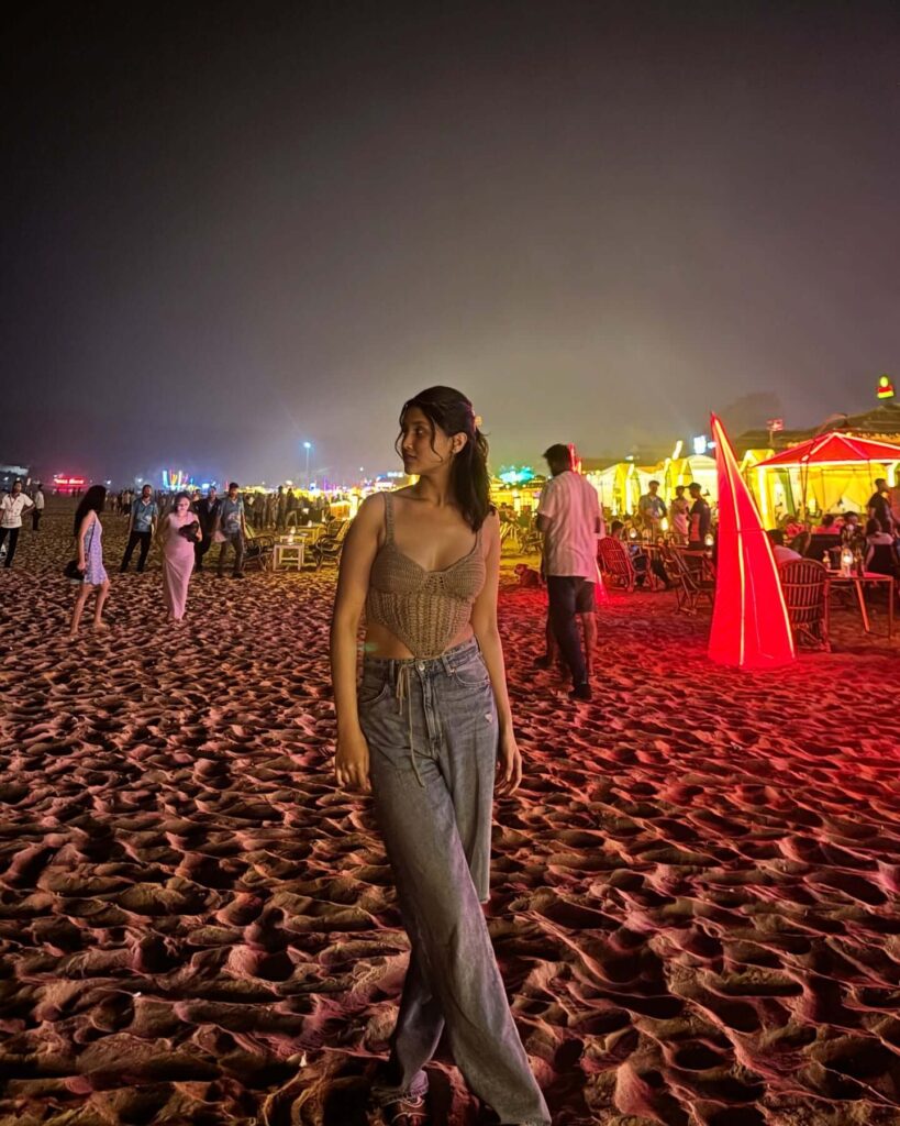 A crowded beach scene at sunset in Goa, with many people relaxing and walking along the shore, and several beach shacks and chairs visible under a warm, colorful sky.