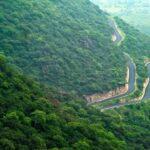 A scenic photograph of a misty, green landscape in the Indian Western Ghats during the monsoon, with rolling hills covered in lush vegetation and a winding road.