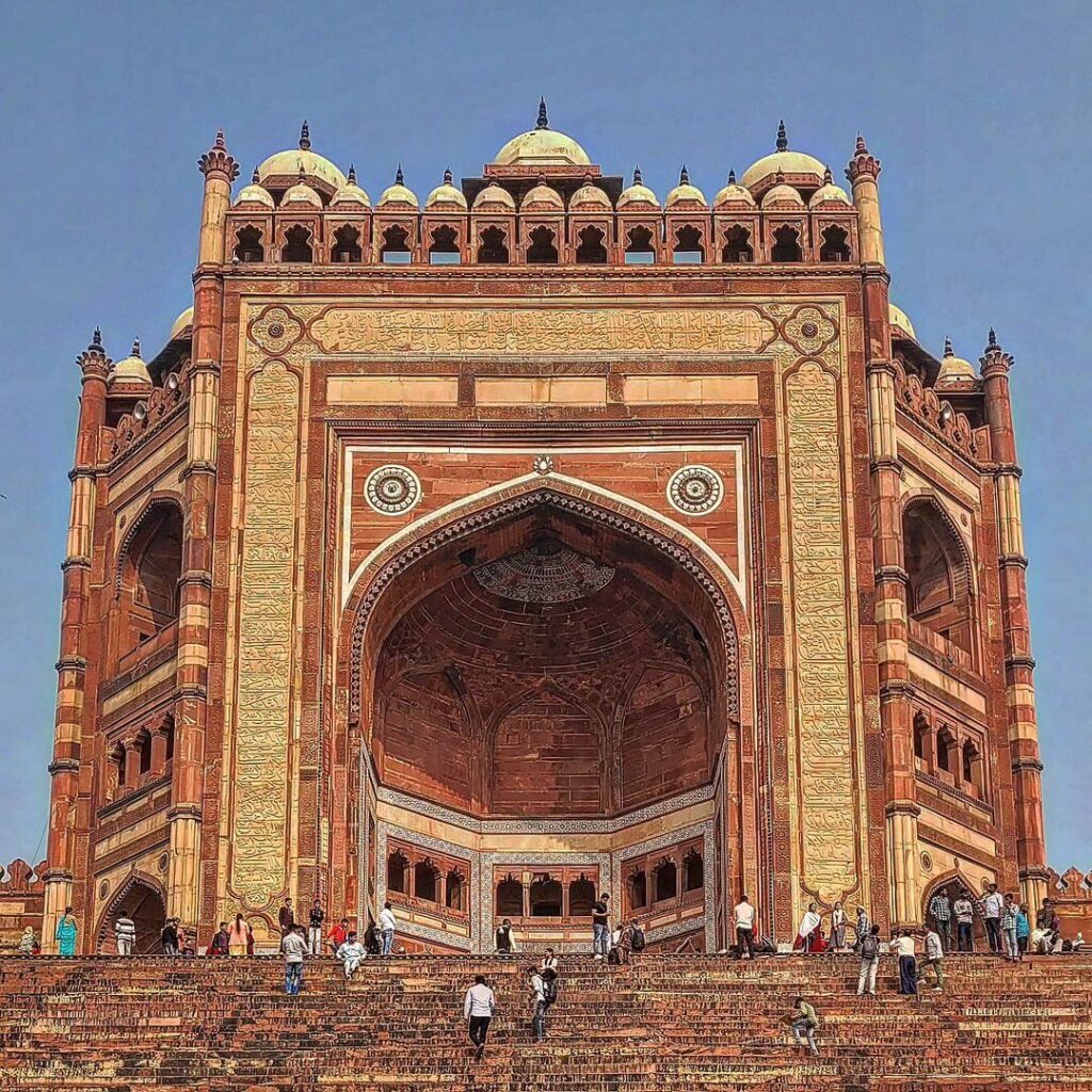 An exterior shot of the Buland Darwaza, the monumental red sandstone gateway to Fatehpur Sikri, with people in the foreground and a clear blue sky.