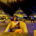 A festive night scene on a beach in Goa during New Year's Eve, with fireworks exploding over the water and a crowd of people watching from the shore.