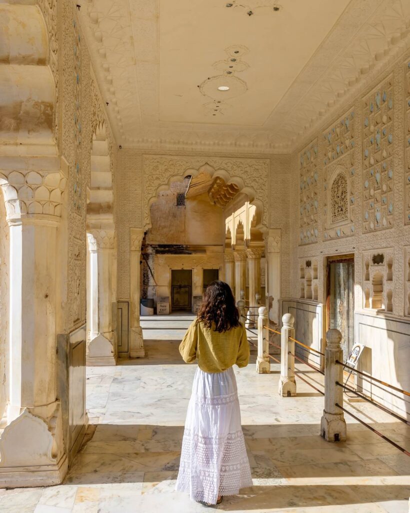 An exterior shot of the Hawa Mahal in Jaipur, a historic red and pink sandstone palace with a honeycomb facade of small windows, standing on a bustling street.