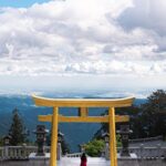 A scenic photograph of a lake with the iconic Mount Fuji in the background, a row of traditional Japanese pagodas, and cherry blossoms in full bloom in the foreground.
