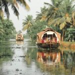 A photograph of a person in a small canoe, paddling through a narrow, tree-lined canal in the Kerala Backwaters.