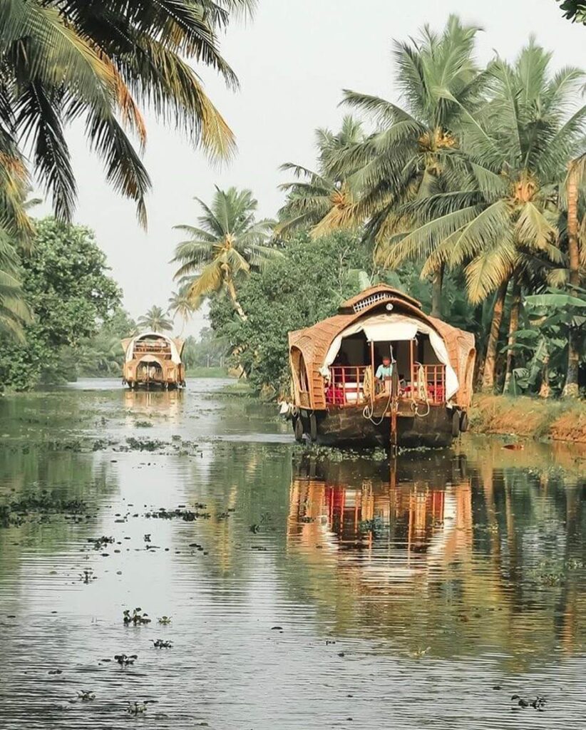 A scenic photograph of a traditional houseboat, or kettuvallam, on a calm backwater channel in Kerala, with lush green palm trees and a tranquil village on the banks.