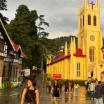 An exterior shot of the Jakhu Temple in Shimla, with a large, vibrant orange statue of Lord Hanuman standing tall against a clear blue sky.