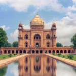 An exterior shot of the Hawa Mahal in Jaipur, a historic red and pink sandstone palace with a honeycomb facade of small windows, standing on a bustling street.