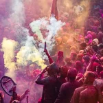 A massive, high-energy crowd of devotees and travelers completely covered in pink and purple powder during the traditional Braj Holi celebration, featuring the best package by Chalotravellers.