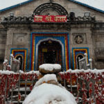 "The ancient stone structure of Kedarnath Temple in Uttarakhand, India, surrounded by snow-capped Himalayan peaks under a clear blue sky, capturing the spiritual essence of the 2026 Yatra opening."