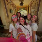 Five young women covered in pink and green colored powder take a joyous group selfie in a traditionally decorated hallway during the Famous Holi Celebration of Pushkar.