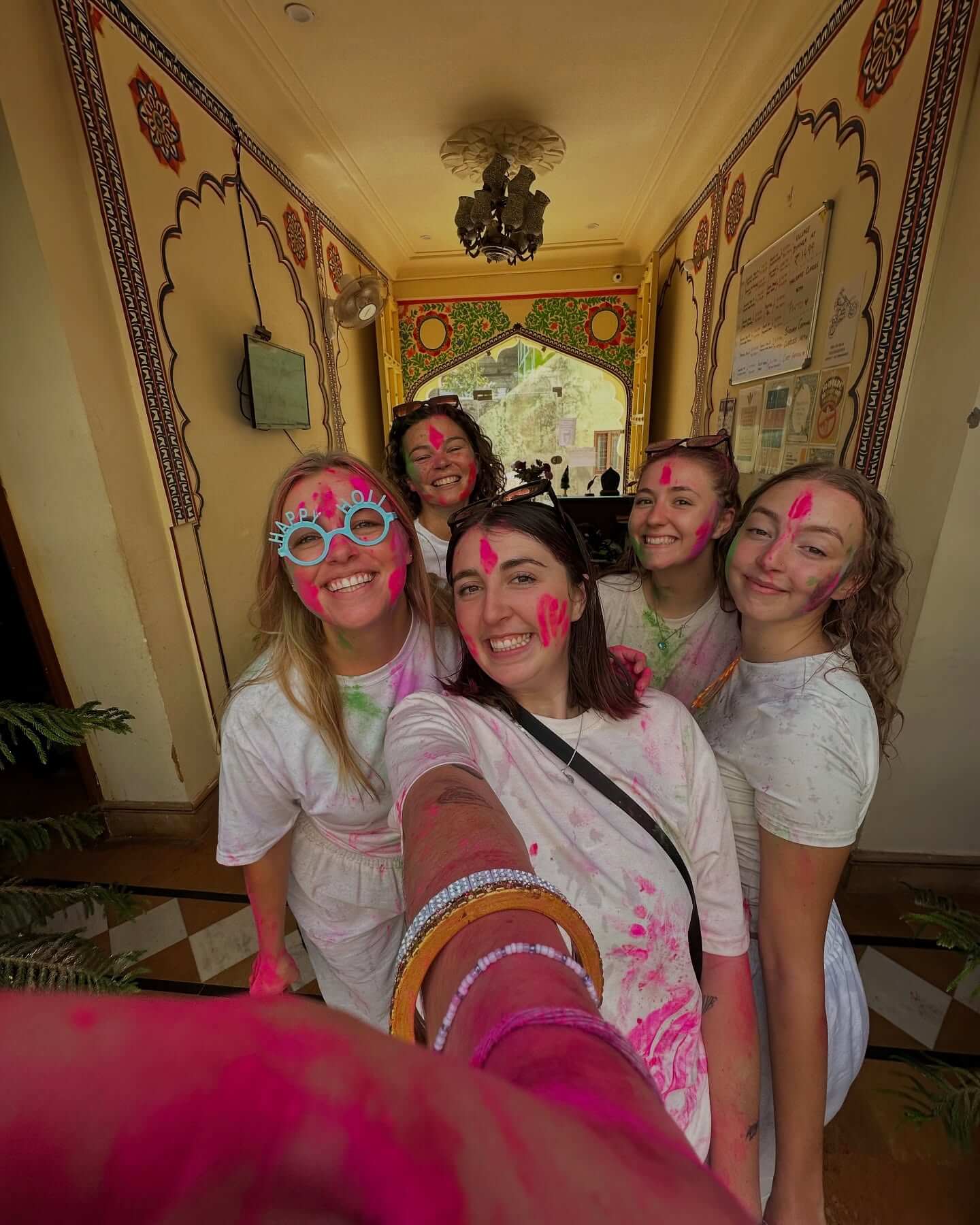 Five young women covered in pink and green colored powder take a joyous group selfie in a traditionally decorated hallway during the Famous Holi Celebration of Pushkar.