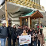 A group of young pilgrims posing with a "Chalo Travellers" banner in front of the Amrit Villa guest house, dressed in heavy winter jackets and beanies. Republic Day LongWeekend Group Trip From Delhi