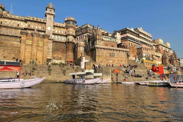 Wooden boat riding past historic ghats and ancient stone palaces in Varanasi along the River Ganges at golden sunrise